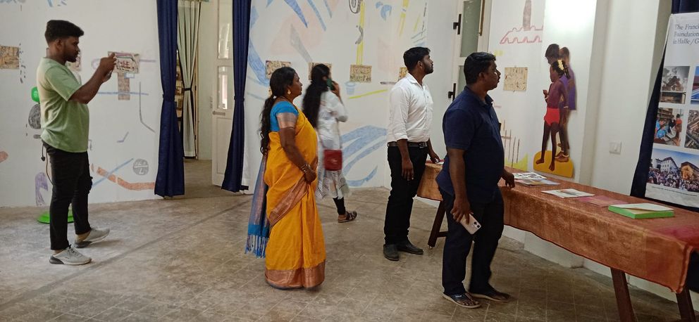 Vier Männer und eine Frau in einem leuchtend orangenem Sari schauen sich eine Kunstausstellung im Ziegenbalg-Haus in Tharangambadi an. 