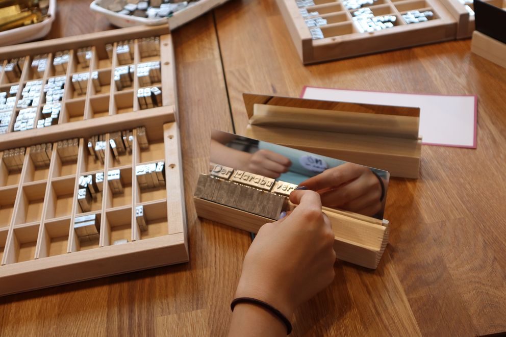 A hand holding a printing block next to a type case filled with movable metal letters used for letterpress printing.