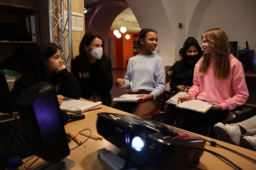 In der Medienwerkstatt im Krokoseum sitzen vier junge Redakteurinnen mit der pädagogischen Fachkraft Frau Hofmann an einem Tisch. Gemeinsam beraten sie sich, während die Kinder mit Klemmbrettern Notizen machen. Im Vordergrund ist ein laufender Beamer zu sehen.