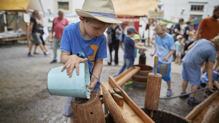 Ein Kind gießt Wasser aus einem Eimer in die Holzrinnen des Wasserspielplatzes auf dem Lindenblütenfest.