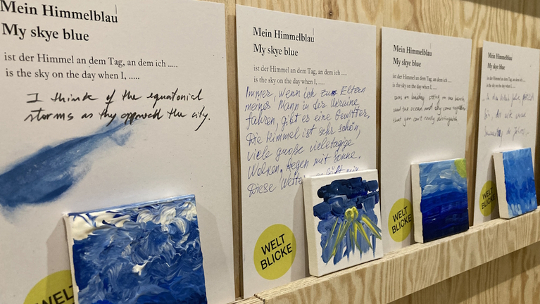 Blick in die Ausstellung mit dem von den Besucherinnen der Ausstellung »Weltblicke« gemischten Himmelblau auf einer Leinwand und der handschriftlichen Notiz zum Himmel, an den sie gedacht haben.
