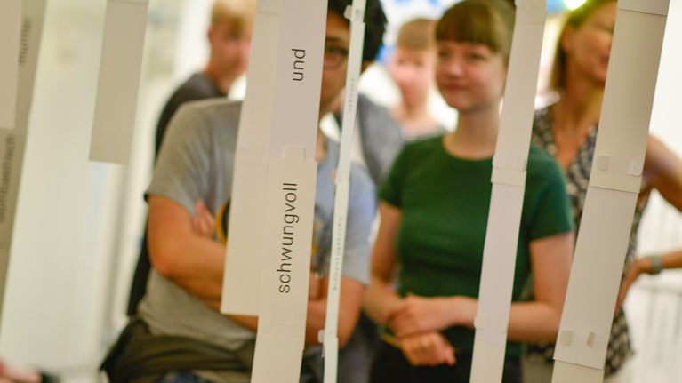 Jugendliche stehen hinter Wortstreifen die in der Ausstellung von der Decke hängen