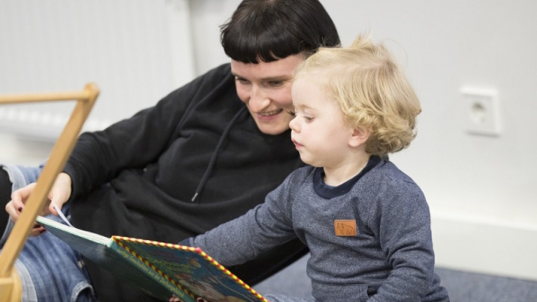 Ein Bild von einer jungen Frau, die seitlich auf dem Teppich liegt und einem kleinen Mädchen aus einem Kinderbuch vorliest. Sie schauen sich gemeinsam die Bilder zu der Geschichte an.