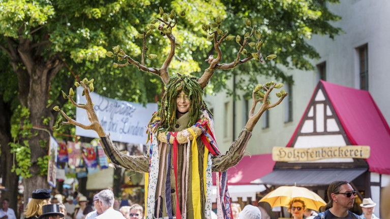 Eine als Baum verkleidete Person steht zwischen den Besucher:innen des Lindenblütenfestes und lächelt in die Kamera