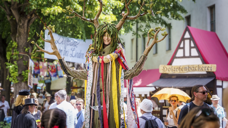 Eine als Baum verkleidete Person steht zwischen den Besucher:innen des Lindenblütenfestes und lächelt in die Kamera
