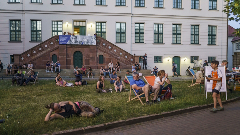 Blick auf die Wiese vor dem Waisenhaus am Abend, Besucher:innen sitzen und unterhalten sich gemütlich