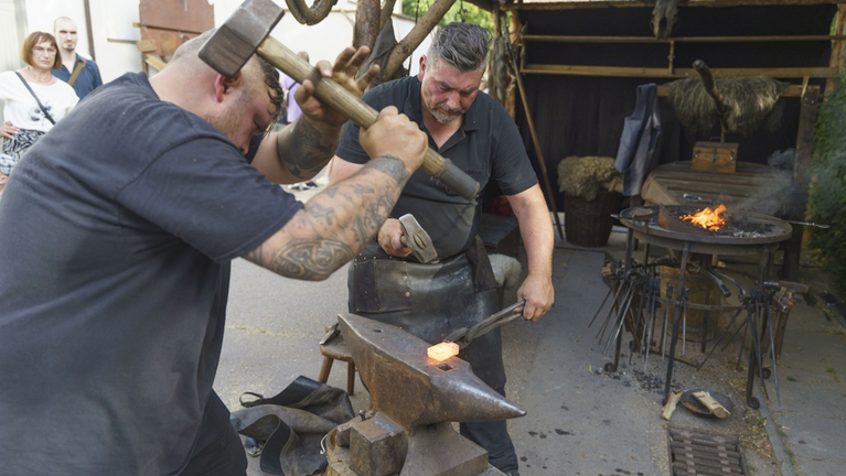 Zwei Schmiede bearbeiten einen glühenden Metallstab mit Hammer und Amboss.