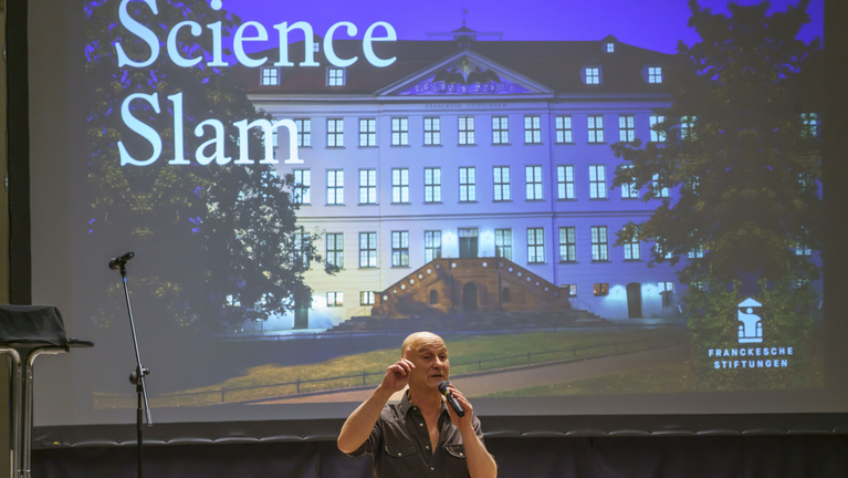 Der Moderator begrüßt zum Science Slam im Freylinghausen-Saal. Hinter ihm die große Leinwand für die Präsentationen der Teilnehmer:innen
