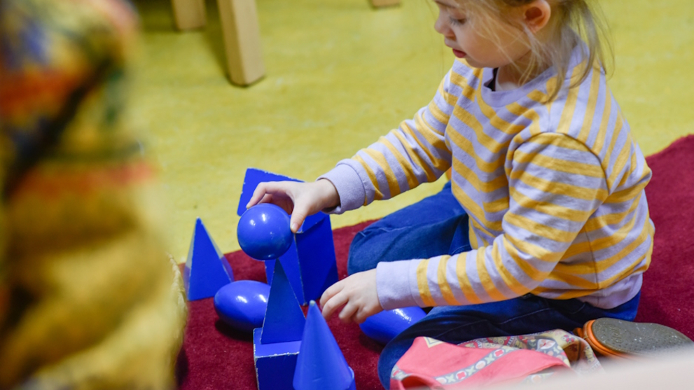 Ein Mädchen spielt auf dem Fußboden mit blauen Kugeln, Kegeln und Quadern aus Holz.